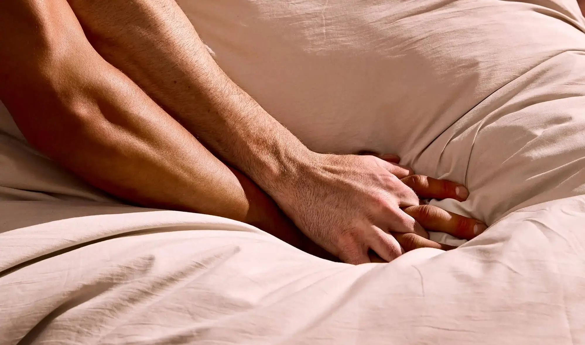 Two men's hands clasped together on bed sheets in golden light