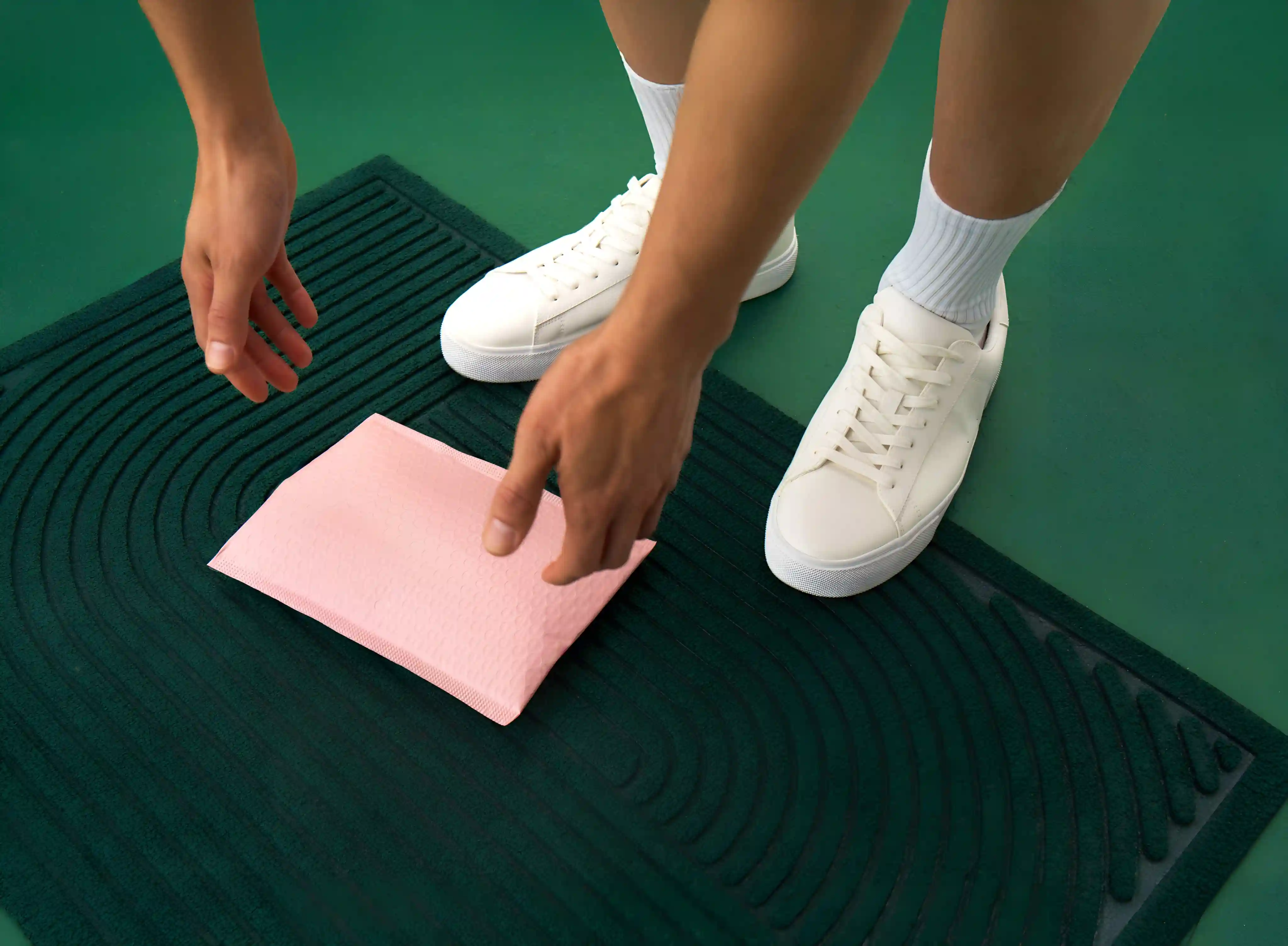 Hands picking up an unlabelled pink delivery package on a green doormat